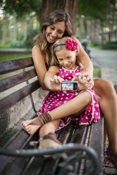 Happy cute little girl and her mother posing for a self portrait at bench in the park. 