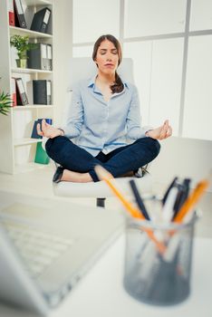 Beautiful young businesswoman practicing yoga in the office. She sits on lotus pose in a chair in front of her desk.