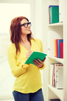 education concept - smiling redhead female student in eyeglasses with textbook in library