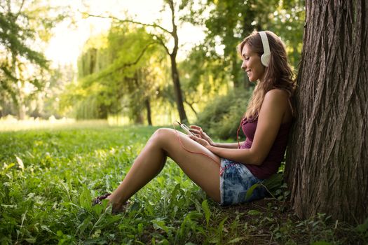 Happy beautiful girl sitting by a tree in a park and listening music from smart phone. 