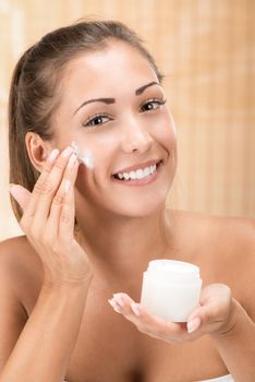 Cute girl preparing to start her day. She is applying moisturizer cream on face. Looking at camera and smiling.
