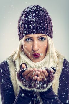 Close-up of beautiful young woman in winter clothes as blowing snowflakes which holds in her hands.