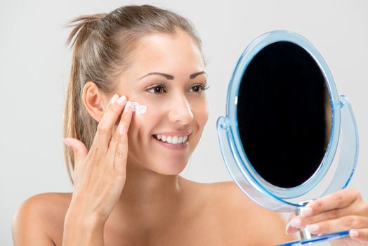 Cute girl preparing to start her day. She is applying moisturizer cream on face. Looking at mirror and smiling. Close-up.