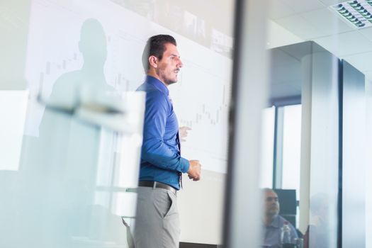 Business man making a presentation in front of whiteboard. Business executive delivering a presentation to his colleagues during meeting or in-house business training. View through glass.