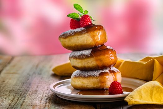 A stack of tree delicious german doughnuts powdered with sugar decorated with raspberry and raspberry sauce on white plate and yellow napkin, blurred blowers as background