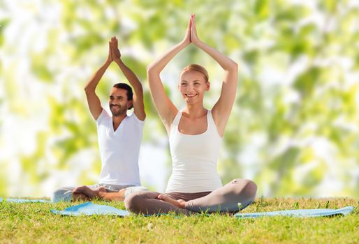 sport, fitness, yoga and people concept - smiling couple meditating and sitting on mats with raised hands over green tree leaves background