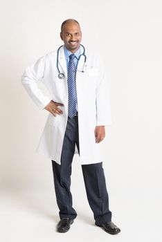 Full length mature Indian male medical doctor in uniform standing on plain background with shadow.