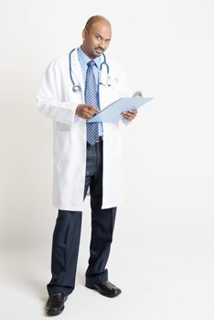 Full length mature Indian male medical doctor in uniform reading on medical report, standing on plain background with shadow.