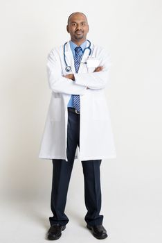 Full length confident mature Indian male medical doctor in uniform looking at camera, standing on plain background with shadow.