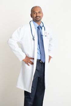 Portrait of confident mature Indian male medical doctor in uniform standing on plain background with shadow.