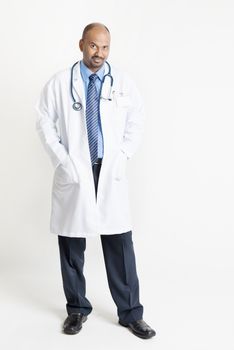 Full length confident mature Indian male medical doctor in uniform standing on plain background with shadow.