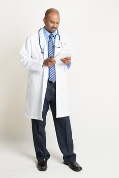 Full length mature Indian male medical doctor in uniform using digital tablet pc, standing on plain background with shadow.