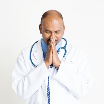 Portrait of mature Indian male medical doctor in uniform praying, standing on plain background with shadow.