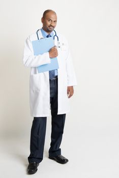 Full length mature Indian male medical doctor in uniform holding medical report folder, standing on plain background with shadow.
