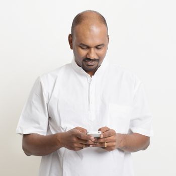 Portrait of mature casual business Indian man using smartphone, mobile apps concept, standing on plain background with shadow.