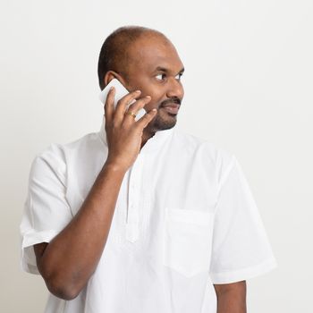 Portrait of mature casual business Indian man calling on smartphone, standing on plain background with shadow.