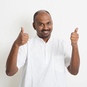 Portrait of happy mature casual business Indian man thumbs up, standing on plain background with shadow.