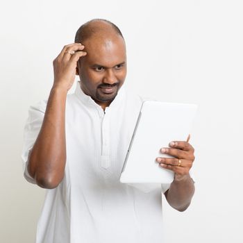 Portrait of mature Indian man headache while reading on digital tablet computer, on plain background with shadow.
