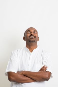 Portrait of mature casual business Indian man arms crossed looking up and smiling, copy space on top, standing on plain background with shadow.