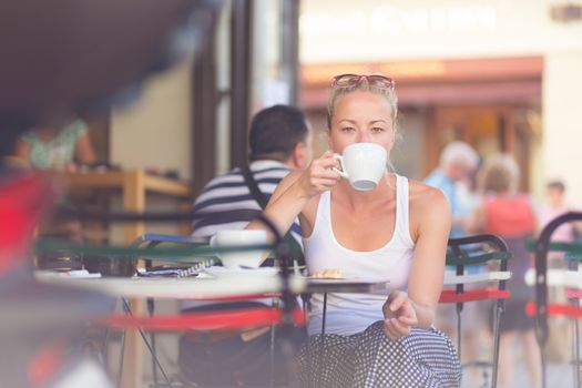 Calm casual blond lady enjoying cup of coffee outdoor in typical italian  street coffee house on warm summer day.