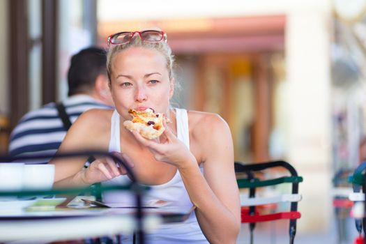 Casual blond lady eating pizza slice outdoor in typical italian street restaurant on hot summer day. Traditional italian fast food eatery.