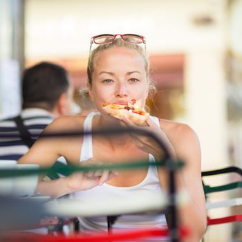 Casual blond lady eating pizza slice outdoor in typical italian street restaurant on hot summer day. Traditional italian fast food eatery.