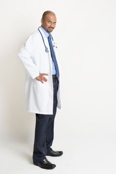 Side view full length confident mature Indian male medical doctor in uniform looking at camera, standing on plain background with shadow.