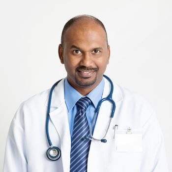 Portrait of mature Indian male medical doctor in uniform smiling, standing on plain background with shadow.