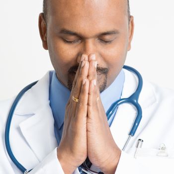 Close up portrait of mature Indian male medical doctor in uniform praying, standing on plain background with shadow.