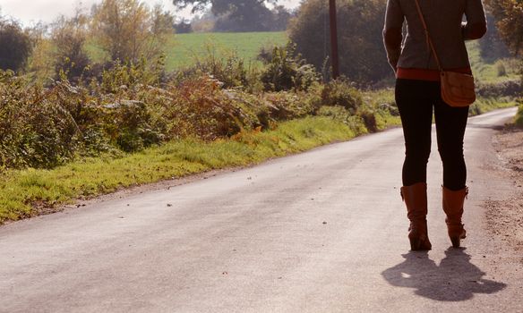 Fashionably dressed young woman walks up a country lane alone in autumn, heading into the sun