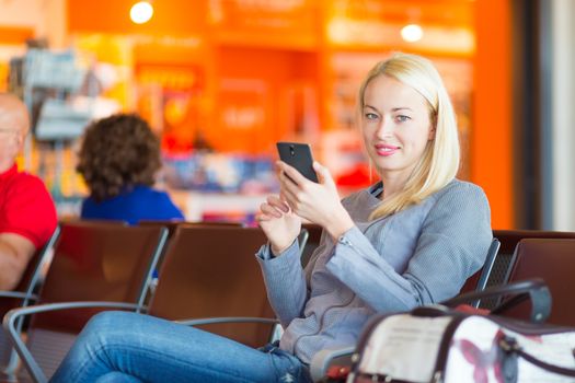 Casual blond young woman using her cell phone while waiting to board a plane at the departure gates. Wireless network hotspot enabling people to access internet conection. Public transport.