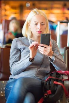 Casual blond young woman using her cell phone while waiting to board a plane at the departure gates. Wireless network hotspot enabling people to access internet conection. Public transport.