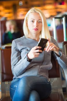 Casual blond young woman using her cell phone while waiting to board a plane at the departure gates. Wireless network hotspot enabling people to access internet conection. Public transport.