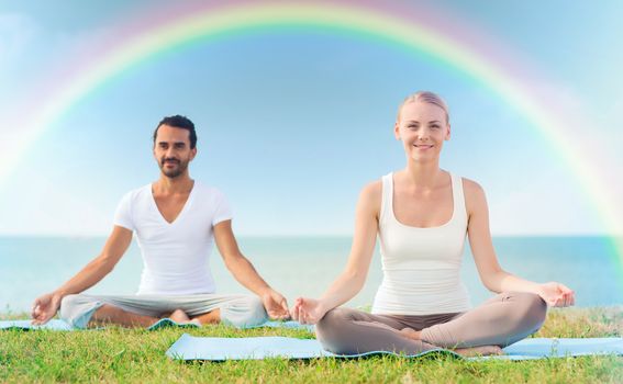 sport, fitness, yoga and people concept - smiling couple meditating and sitting on mats over sea and rainbow in blue sky background