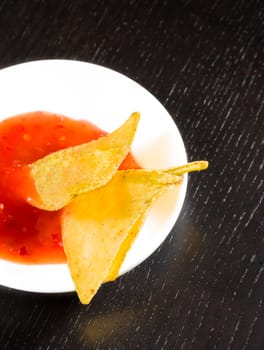 top of view of crisp corn nachos with spicy hot tomato sauce as a snack or appetizer in a white disc on black wood table