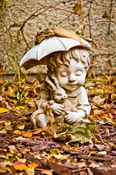 A weathered statue of a young boy holding a rabbit and an umbrella, which is covered in leaves in my backyard.