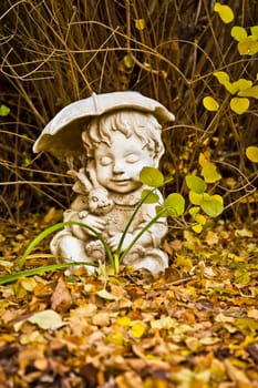 A weathered statue of a young boy holding a stuffed rabbit and an umbrella, surrounded by a carpet of dry leaves in my backyard.