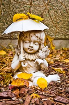 A weathered statue of a young girl holding an umbrella an a tiny bird, surrounded by dry leaves and bark chips in my backyard.