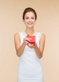 holidays, presents, wedding and happiness concept - smiling woman in white dress holding red gift box over beige background