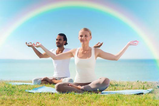 sport, fitness, yoga and people concept - smiling couple meditating and sitting on mats over sea and rainbow in blue sky background