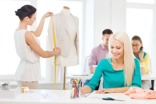 startup, education, fashion and office concept - smiling designers drawing sketches and measuring jacket on mannequin in office