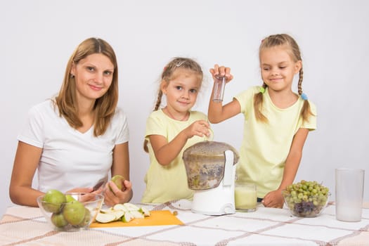 Young beautiful mother and two daughters sitting at a table squeezed juice from pears and grapes with a juicer