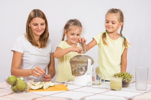 Young beautiful mother and two daughters sitting at a table squeezed juice from pears and grapes with a juicer