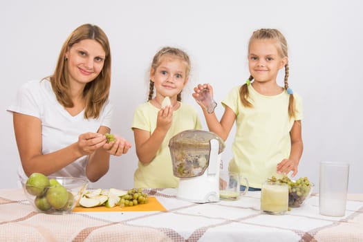 Young beautiful mother and two daughters sitting at a table squeezed juice from pears and grapes with a juicer