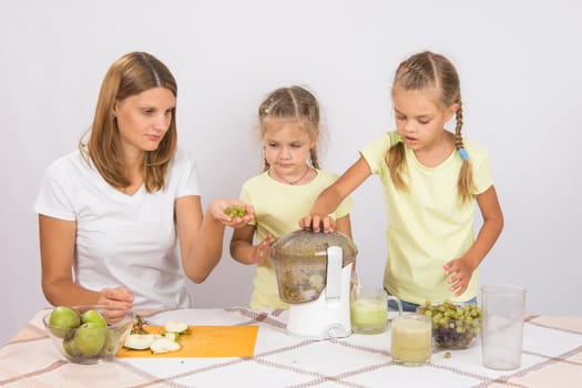 Young beautiful mother and two daughters sitting at a table squeezed juice from pears and grapes with a juicer