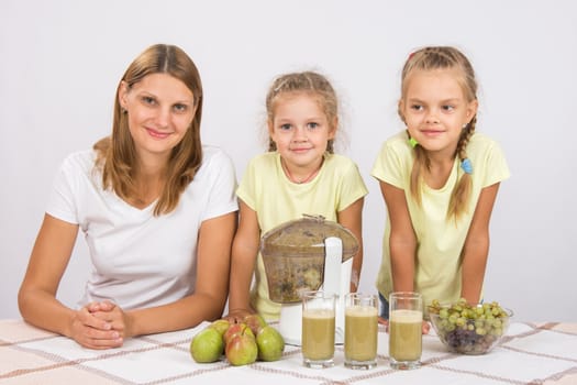Young beautiful mother and two daughters sitting at a table squeezed juice from pears and grapes with a juicer