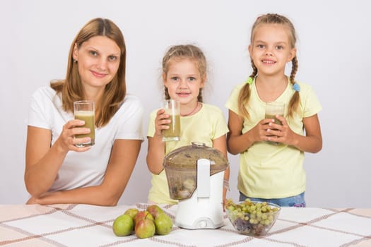 Young beautiful mother and two daughters sitting at a table squeezed juice from pears and grapes with a juicer