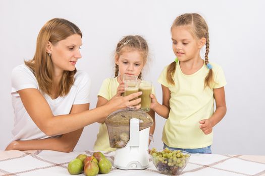 Young beautiful mother and two daughters sitting at a table squeezed juice from pears and grapes with a juicer