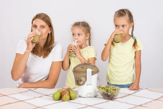 Young beautiful mother and two daughters sitting at a table squeezed juice from pears and grapes with a juicer