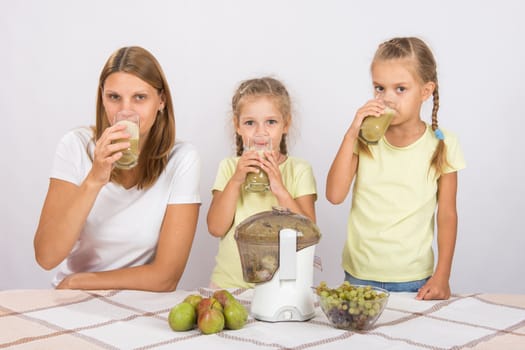 Young beautiful mother and two daughters sitting at a table squeezed juice from pears and grapes with a juicer
